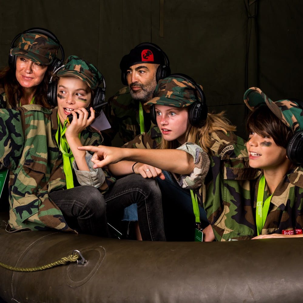 Fire personer i camouflageuniformer og headset, to er børn, en med ansigtsmaling, der peger og bruger en radio i et militært telt.