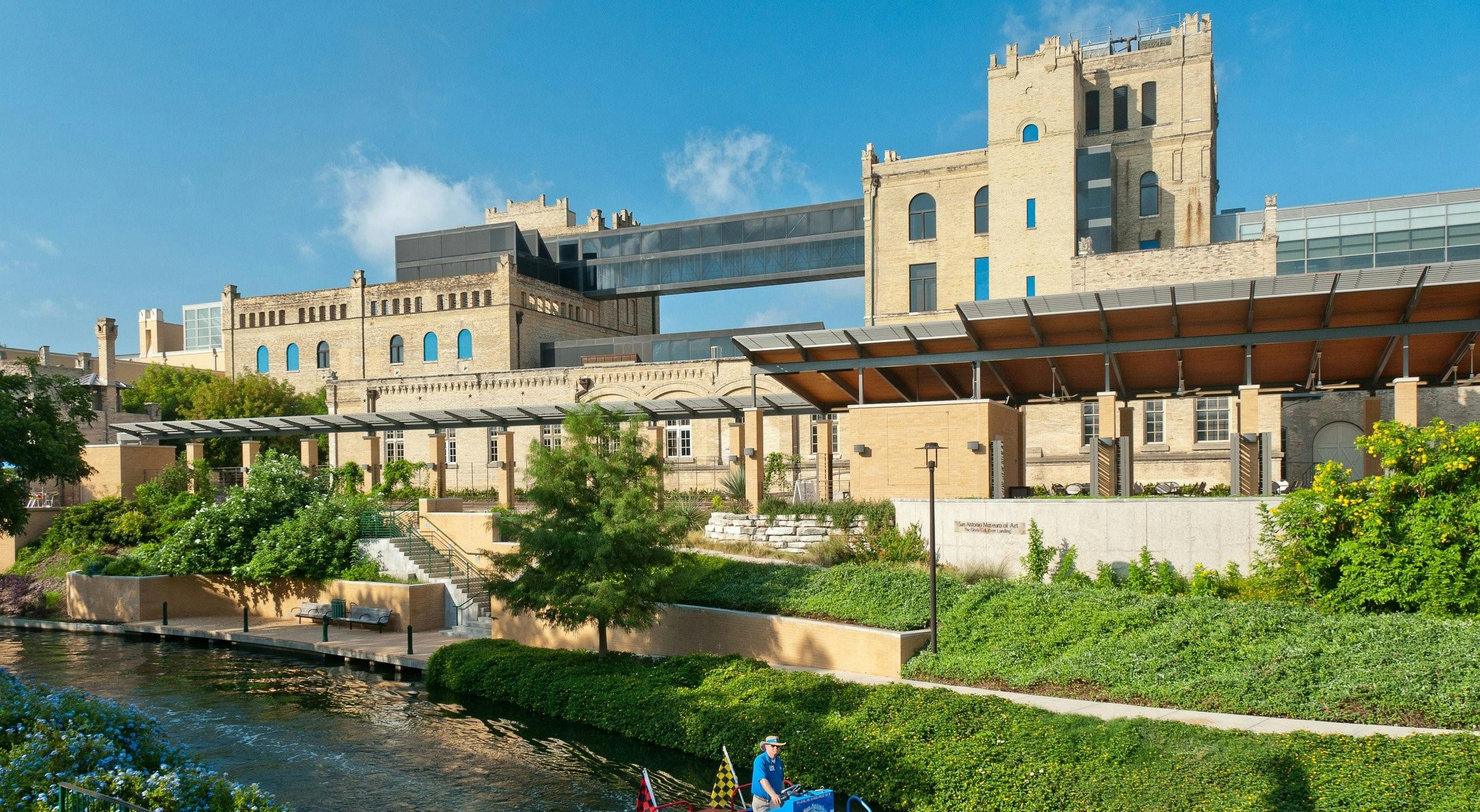 Un edificio storico con un ponte di vetro, circondato da una vegetazione lussureggiante e da un fiume, sotto un cielo azzurro e limpido.