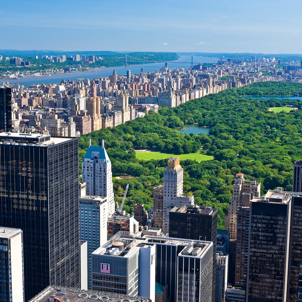 Aerial view of Manhattan with Central Park's greenery on the right, surrounded by tall buildings under a clear blue sky.
