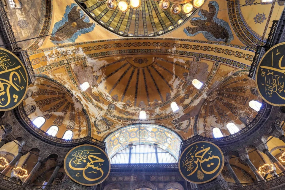 Interior of a grand dome with ornate Islamic calligraphy and intricate mosaics, illuminated by natural light.