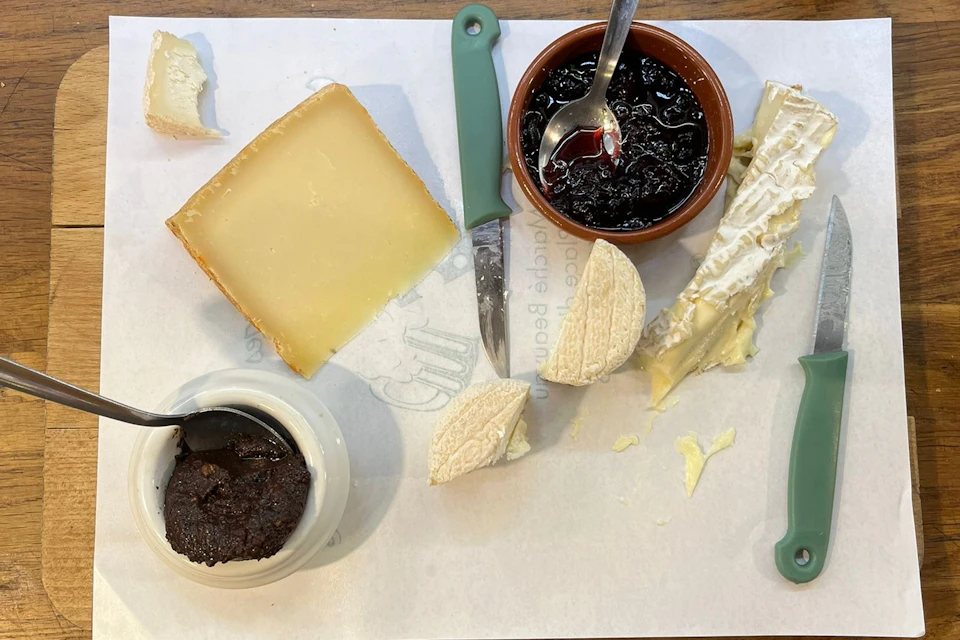 A table laid with a selection of cheeses, a jar of jam, a jar of spread, and knives on a sheet of parchment.
