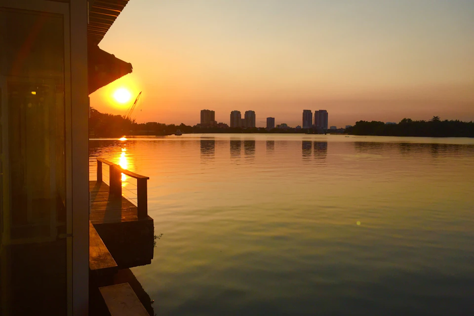 Sunset over a calm lake with buildings on the distant shore, viewed from a wooden deck with railing.