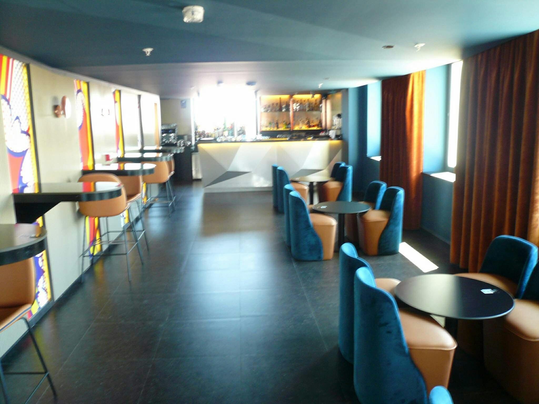 Modern lounge area with round tables and blue chairs, orange bar stools along the wall, a bar at the back, and large windows with curtains.