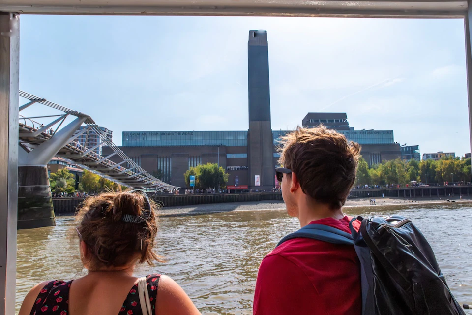 Two people on a boat observe a riverside view of the Tate Modern and the Millennium Bridge in London on a sunny day.