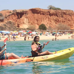 Two people kayaking in the ocean near a crowded beach with colorful umbrellas and a backdrop of red cliffs.
