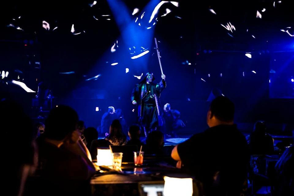 A performer on stage with dramatic lighting and effects, viewed by an audience seated at tables in a dimly lit venue.