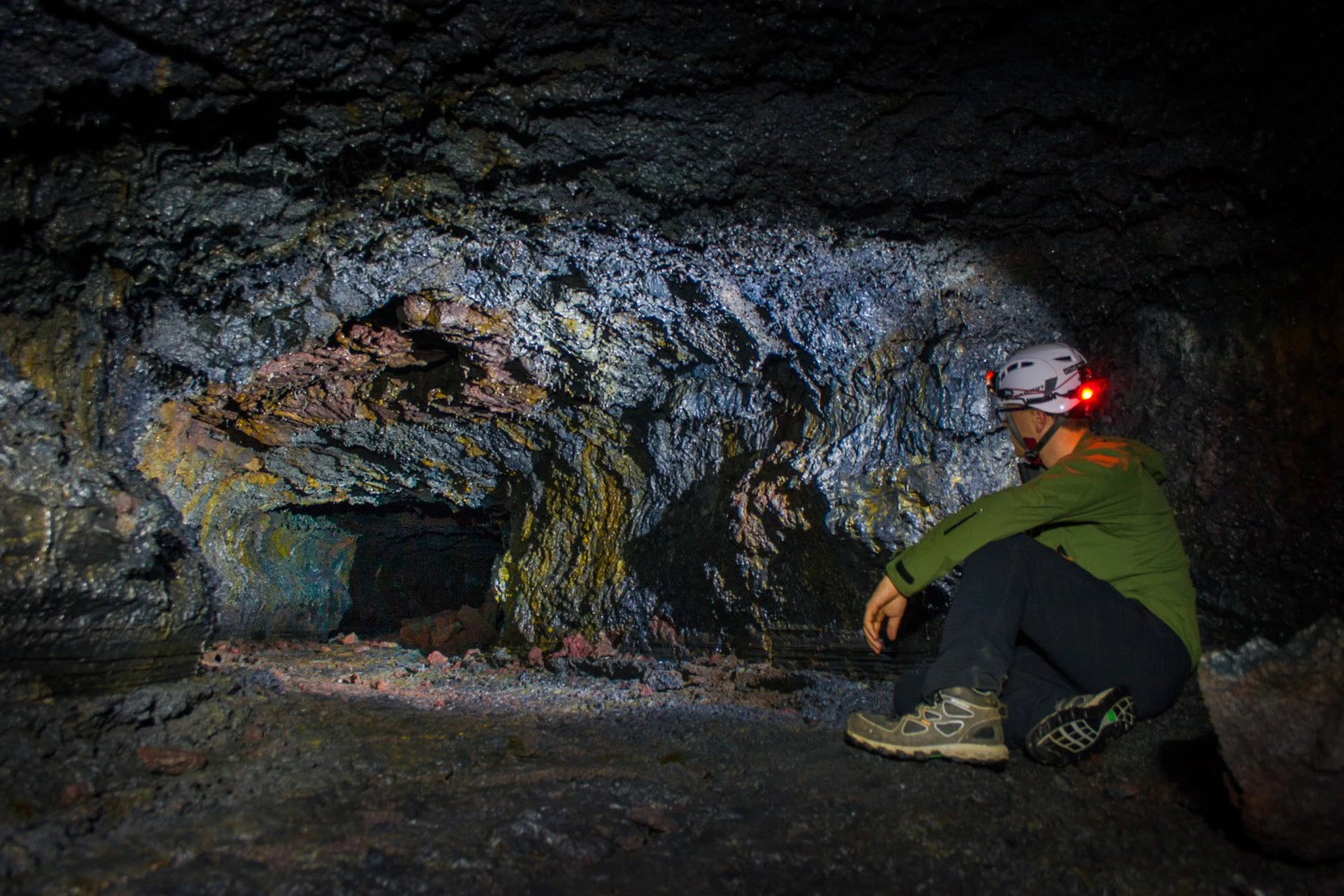 Person wearing a helmet with a headlamp sits inside a dimly lit cave with rugged, rocky walls and a narrow passage.