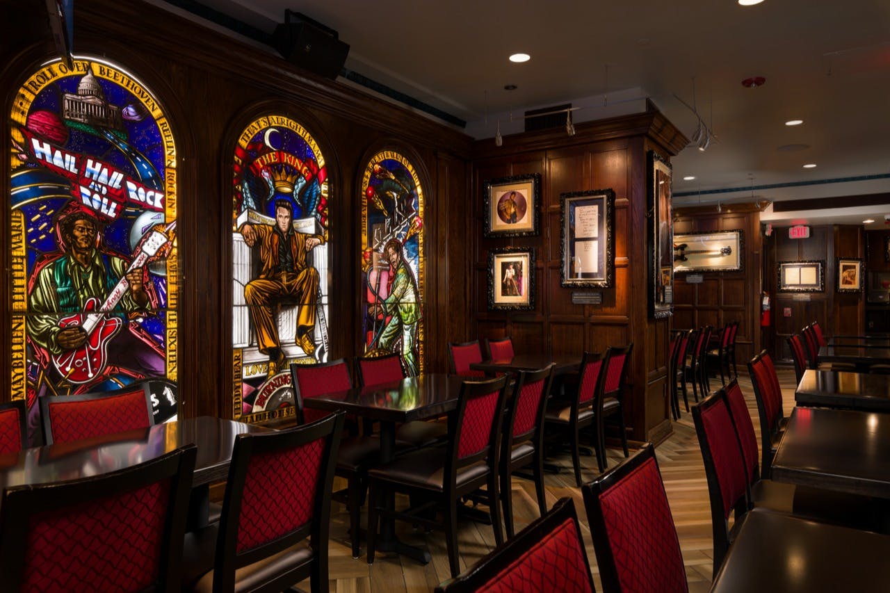 A room with stained glass art depicting musicians, framed pictures on walls, and tables with chairs featuring red upholstery.