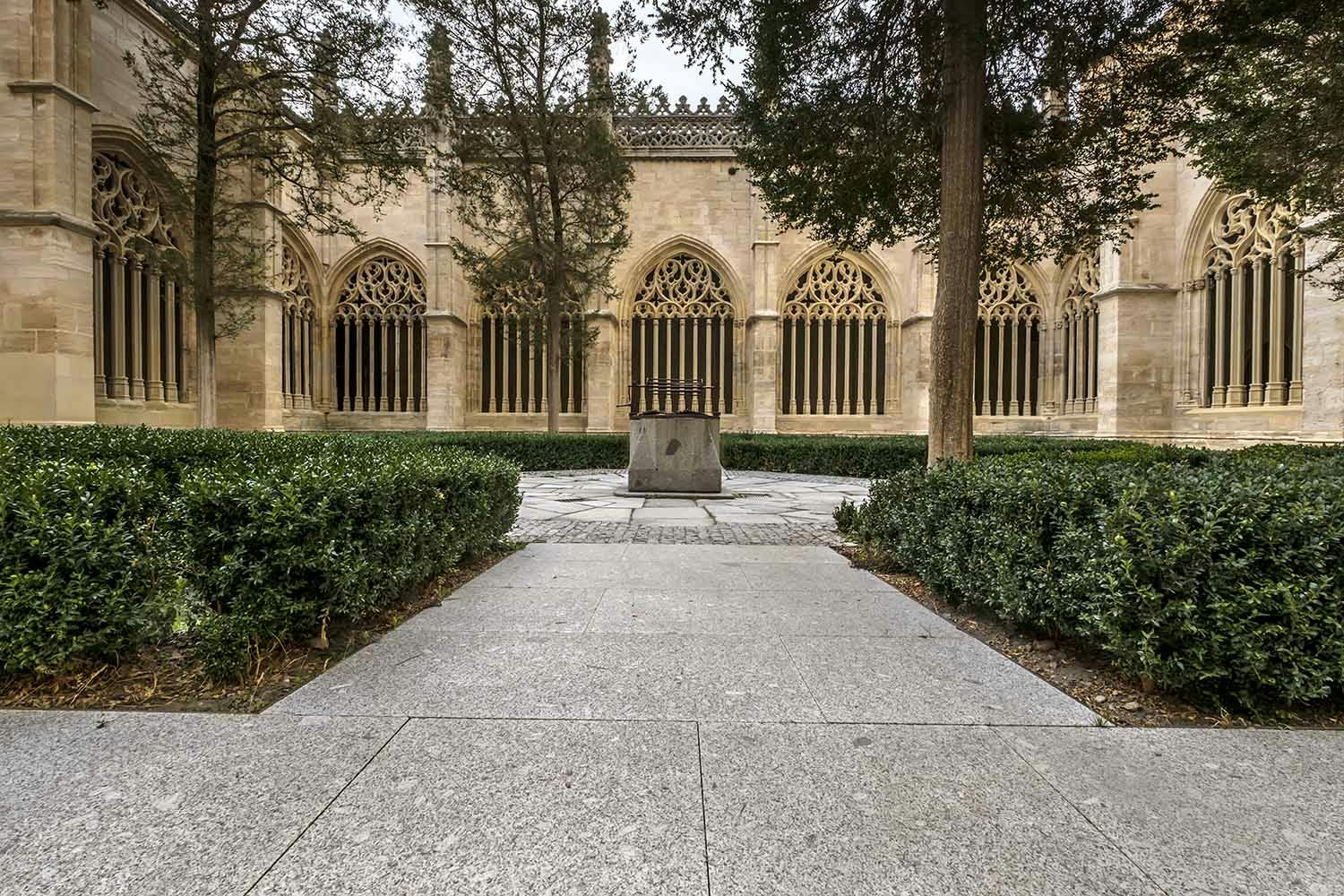Stone walkway leading to a central monument, surrounded by trimmed hedges and trees, in front of a gothic-style building with arched windows.