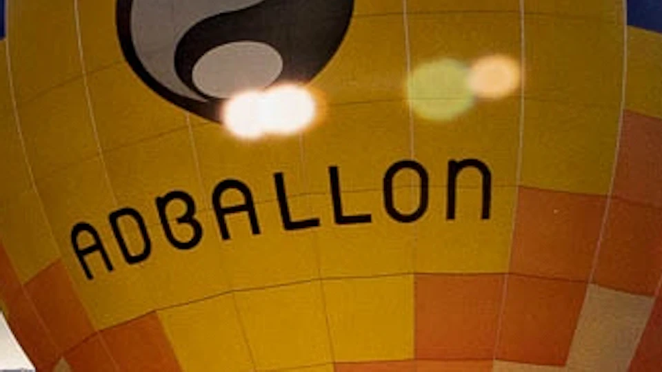 Several colorful hot air balloons on the ground with one flying in the sky under a partly cloudy sky. People and cars nearby.