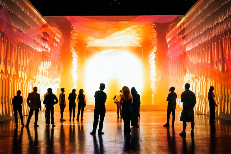 鮮やかなオレンジとイエローのトーンを持つ、明るくカラフルで没入型のアートインスタレーションを観察している人々のシルエット。