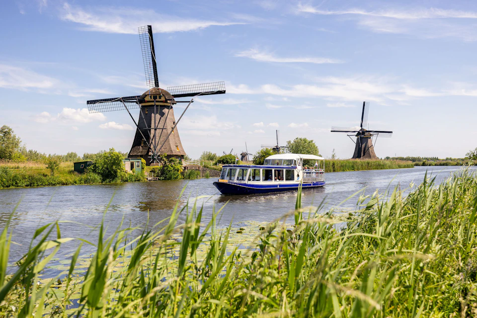 A canal boat passes by two traditional windmills on a sunny day, surrounded by greenery and clear blue sky.