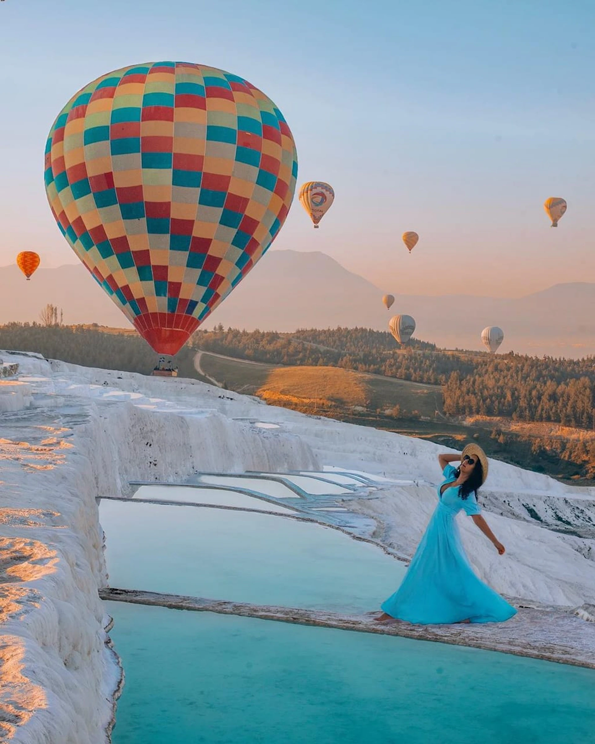 A woman in a blue dress stands by terraced pools with colorful hot air balloons flying over a scenic landscape at sunset.