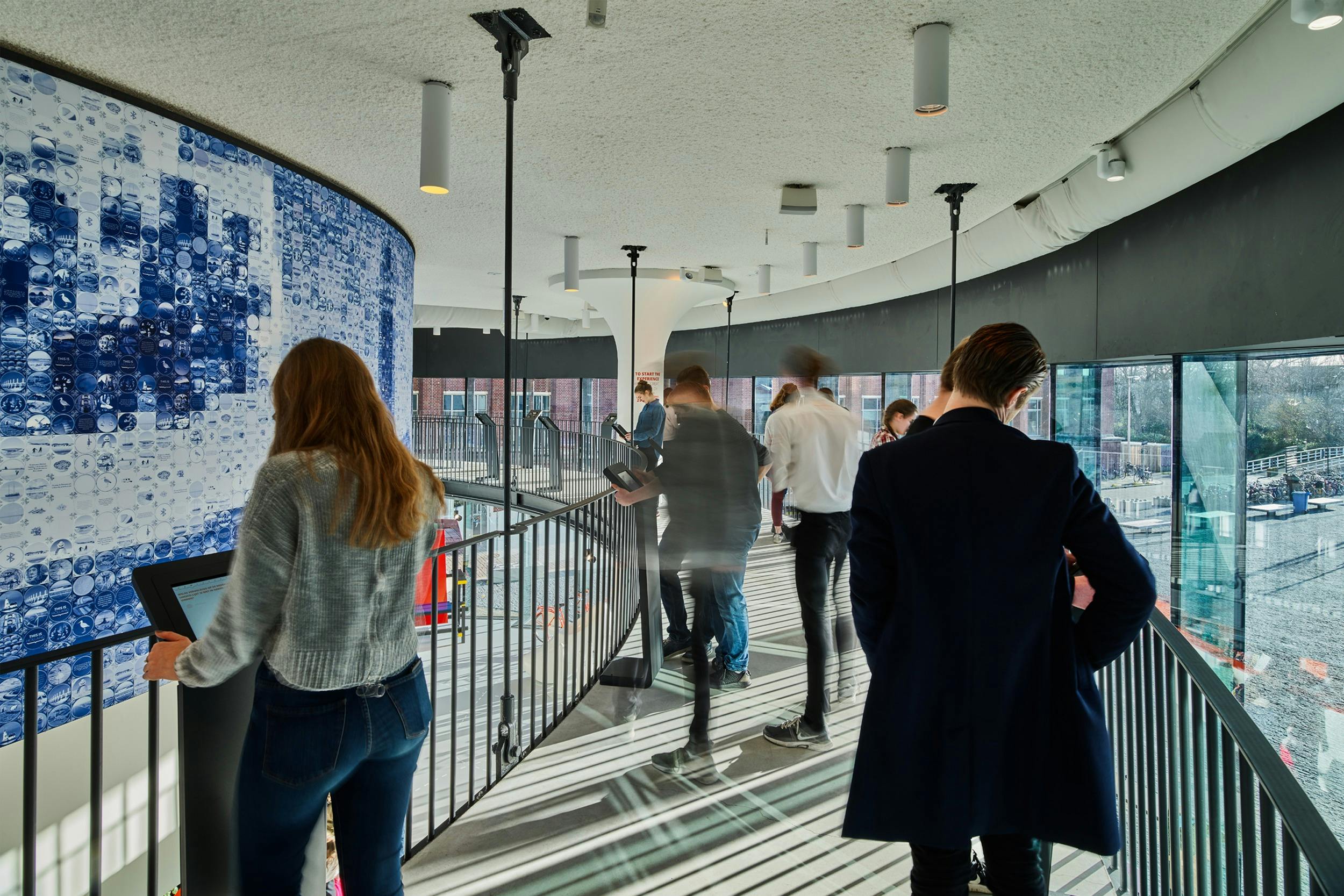 Lidé chodí a mluví v zakřivené chodbě se stěnou s okny, moderním osvětlením a nástěnnou malbou s modrým vzorem.