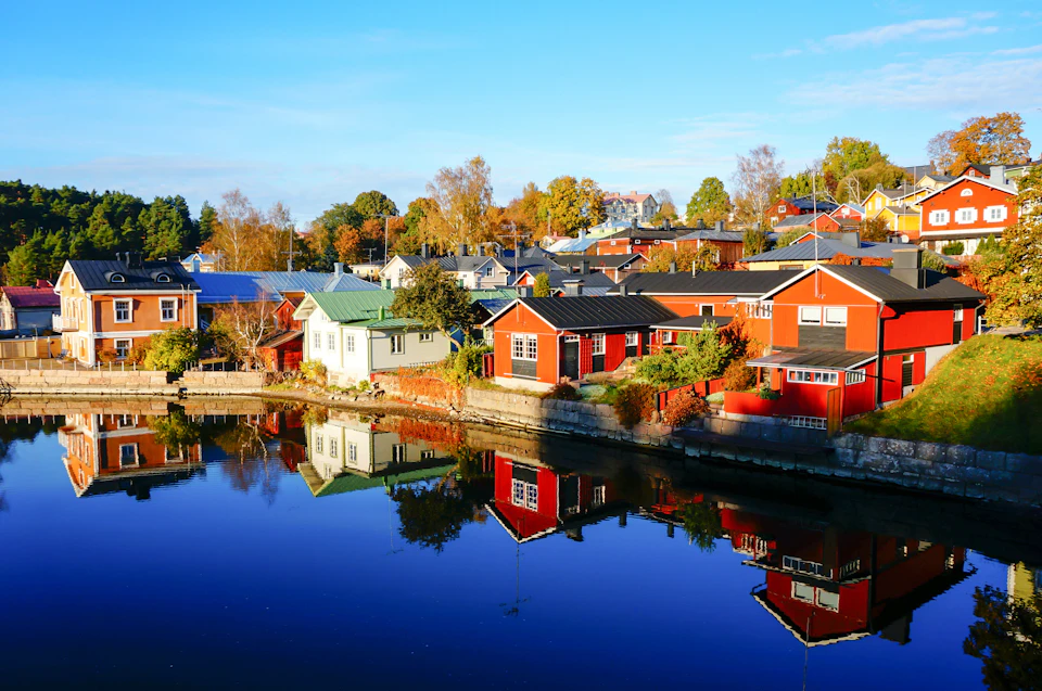 Colorful houses reflected in a calm body of water, with autumn foliage in the background under a clear blue sky.