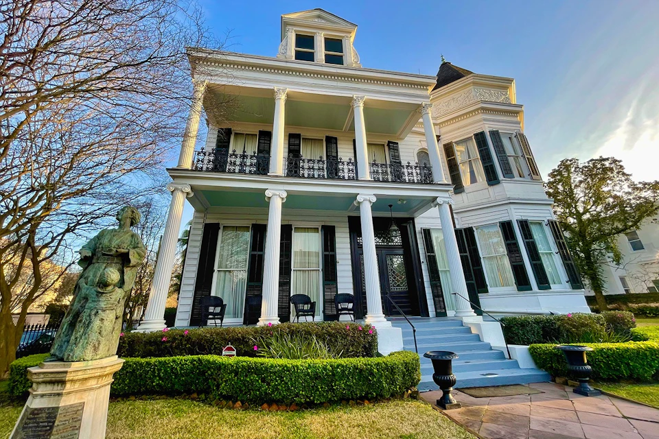 A large, white, colonial-style house with two stories, a balcony, and decorative columns, surrounded by a manicured garden.