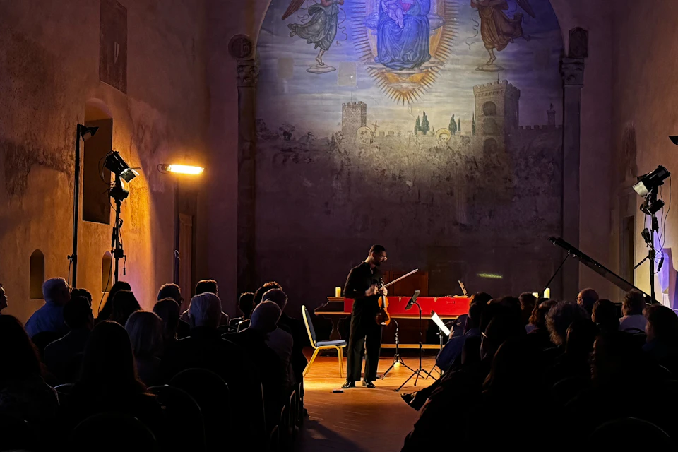 A violinist performs in a dimly lit hall with a large mural on the wall behind them, surrounded by an audience.