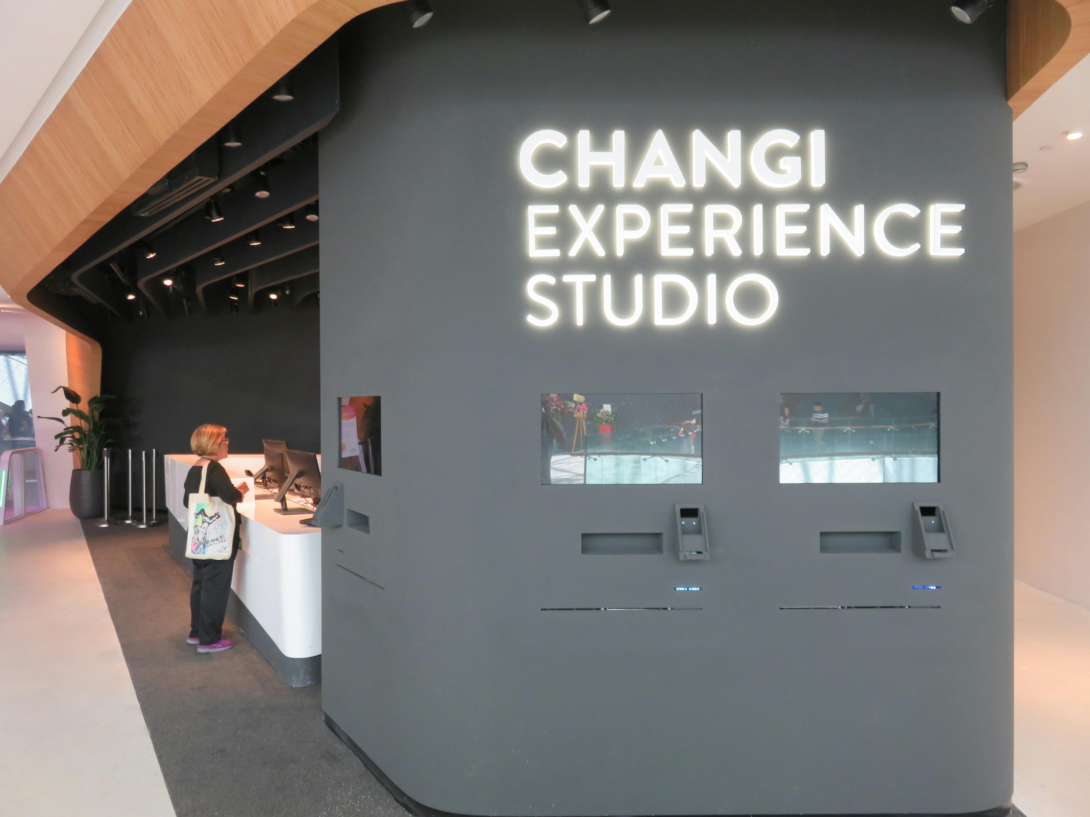U recepce studia Changi Experience Studio stojí osoba s velkou reklamní cedulí a samoobslužnými kiosky.