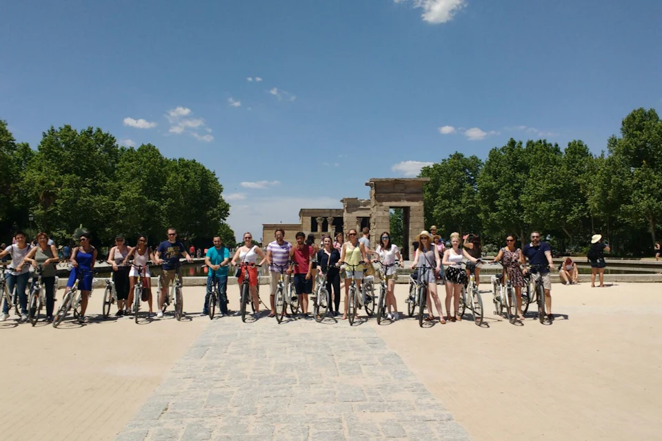 A group of people with bicycles stands in front of an ancient stone building on a sunny day, surrounded by trees.