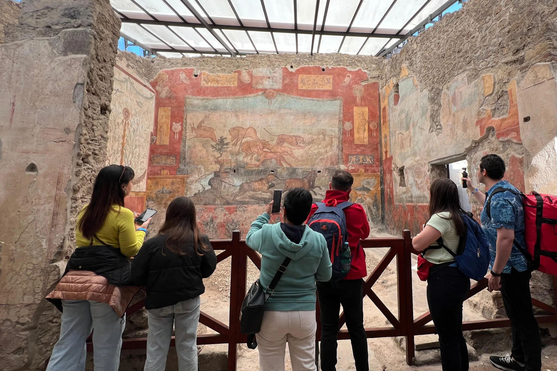 Five people observe and photograph ancient murals on the walls of a historical site under a transparent roof.