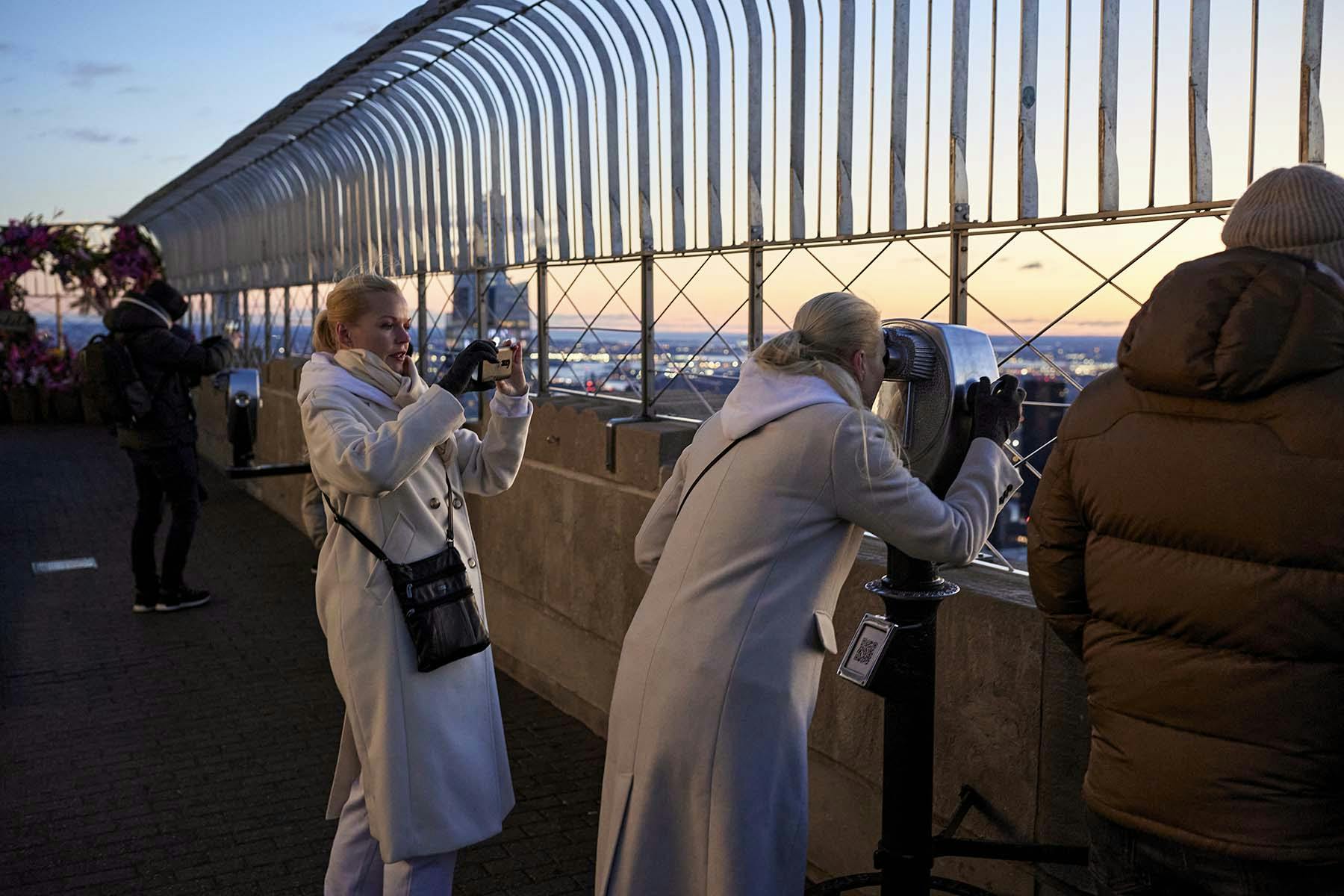 Due donne in camice bianco osservano la città da una piattaforma di osservazione all'aperto con un binocolo e una macchina fotografica.