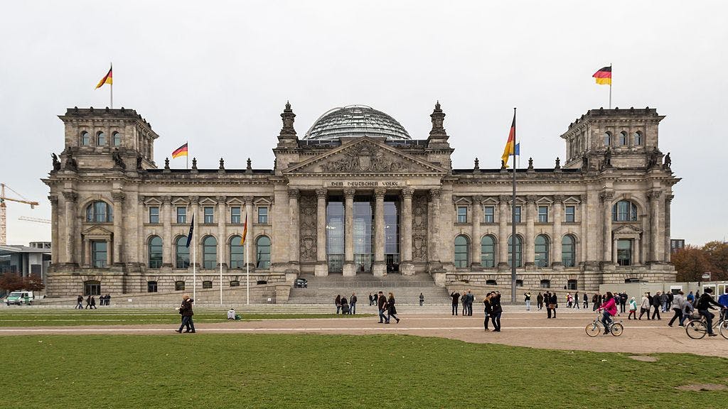 O edifício do Reichstag em Berlim, adornado com bandeiras alemãs. Pessoas caminham e andam de bicicleta em primeiro plano num dia nublado.