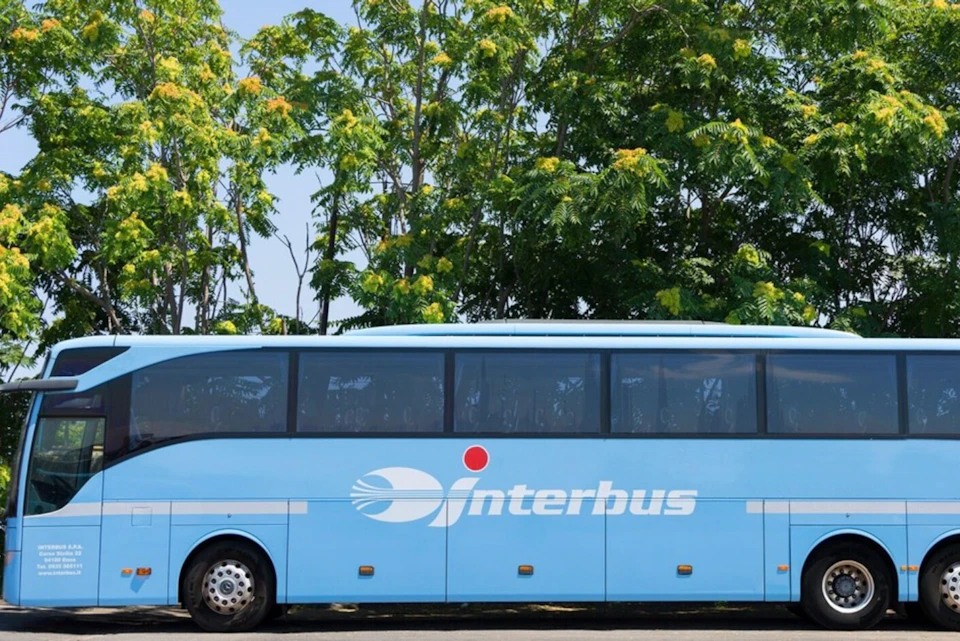 A blue intercity bus with the logo 'Interbus' parked near leafy green trees on a sunny day.