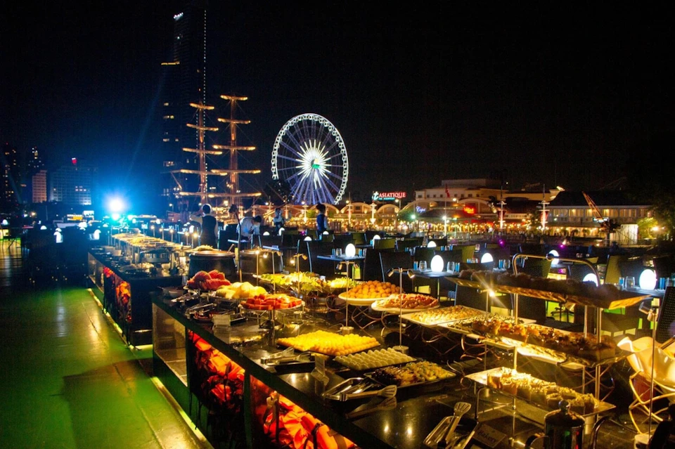 Outdoor night dining setup with a buffet table full of food, people dining, a ferris wheel, a tall ship, and illuminated signs in the background.