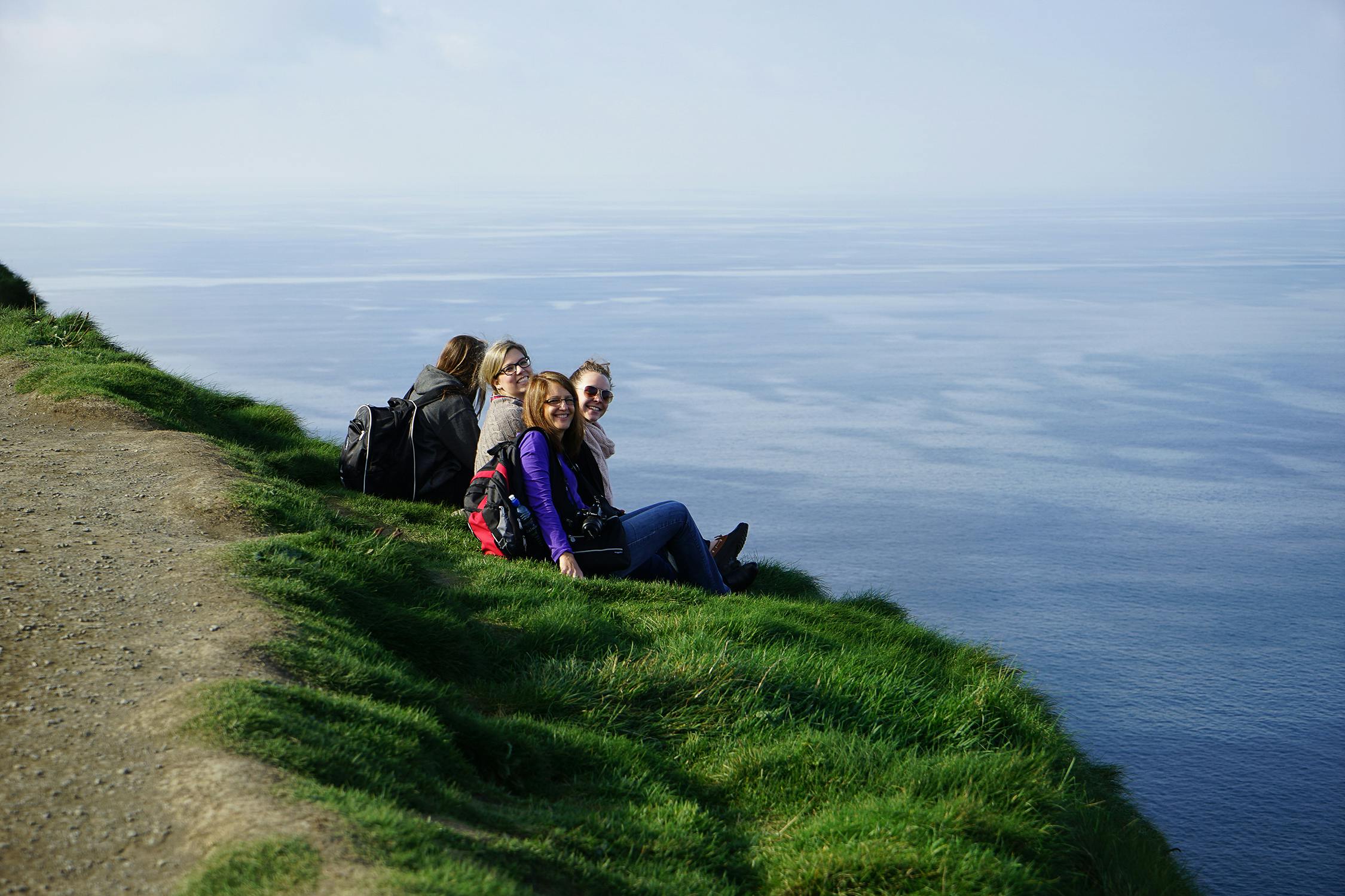 Four people with backpacks sitting on a grassy cliff edge, overlooking a calm body of water under a clear sky.