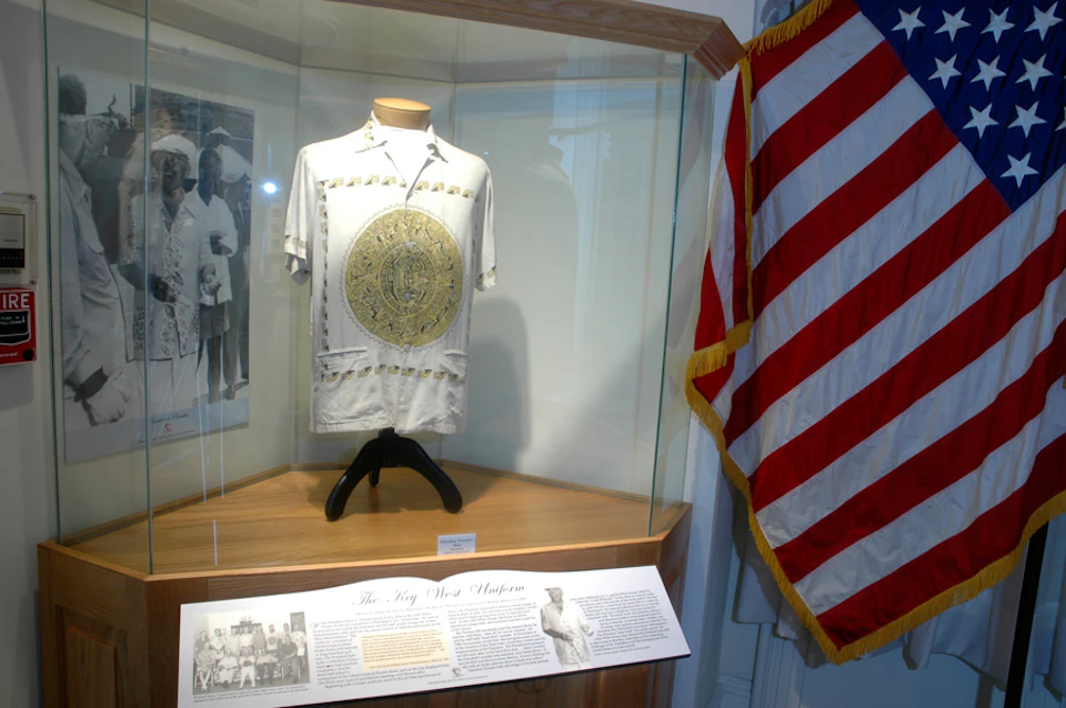 A displayed shirt with a large central emblem in a glass case beside a United States flag and informational text below.