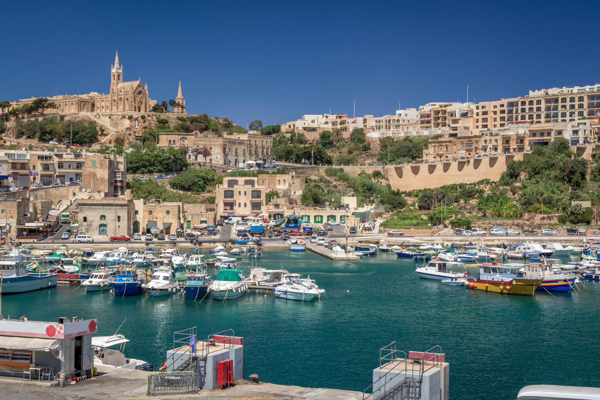 A harbor with various boats docked, surrounded by buildings and a hilltop church under a clear blue sky.