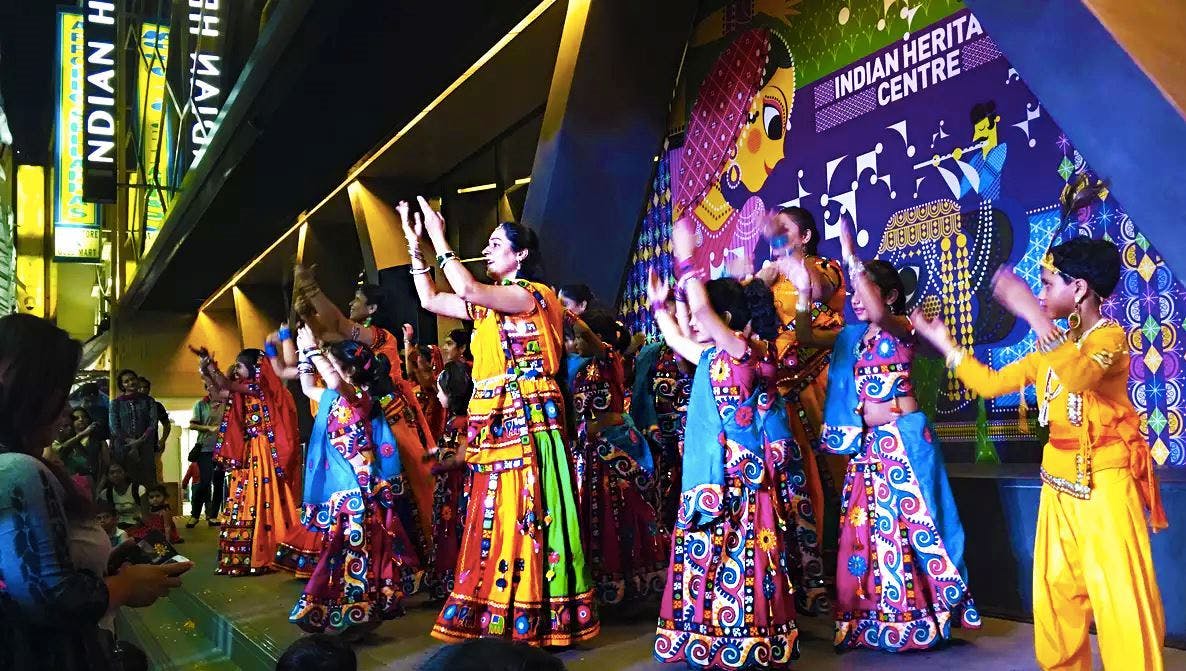 Группа людей в колоритном традиционном наряде энергично танцует на сцене с праздничным декором на заднем плане.