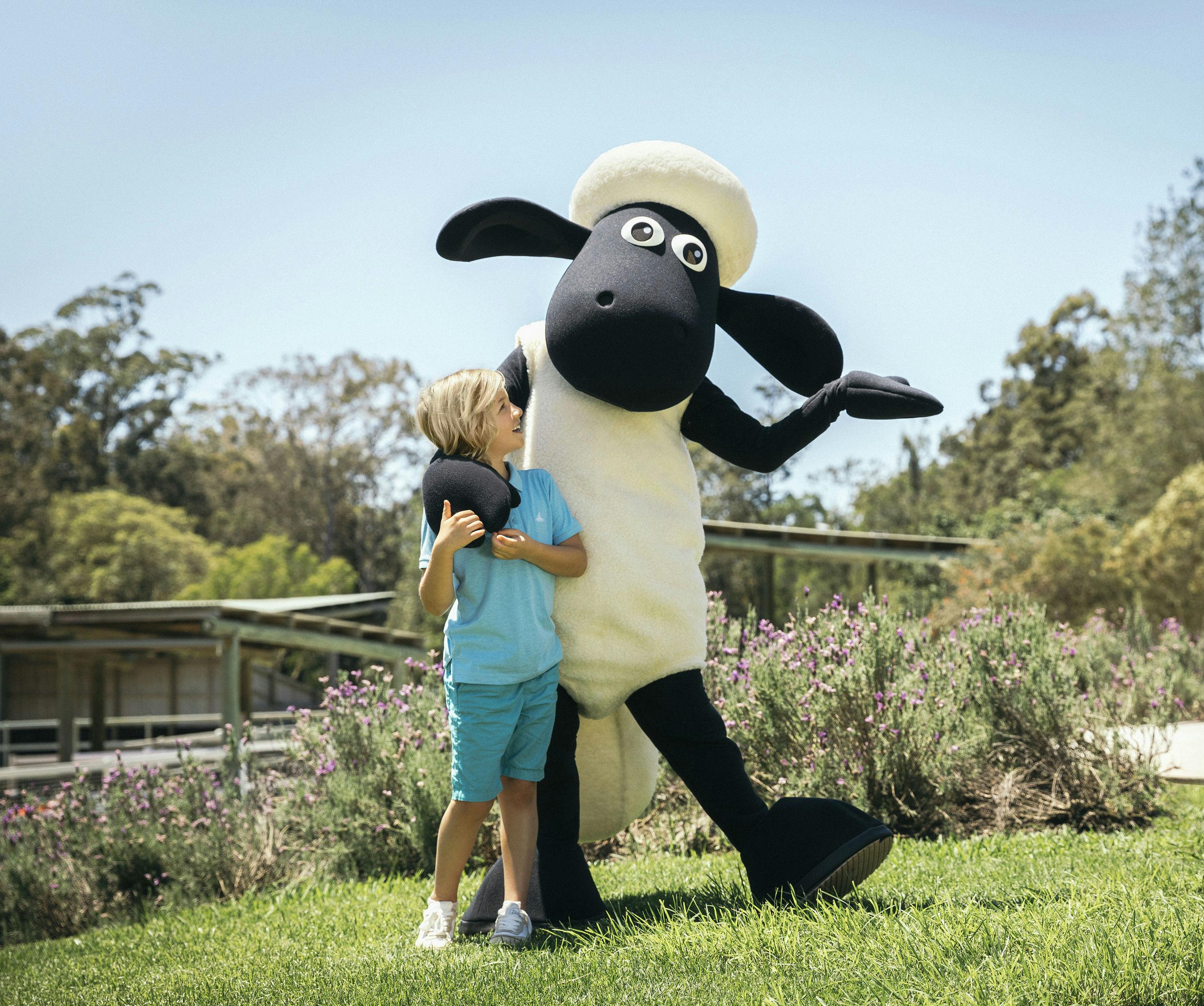 Ένα χαμογελαστό παιδί στα μπλε στέκεται δίπλα σε μια μεγάλη μασκότ προβάτου σε μια καταπράσινη περιοχή με λουλούδια, δέντρα και καθαρό ουρανό στο παρασκήνιο.