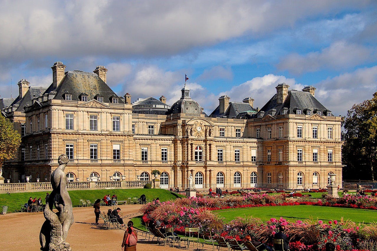 卢森堡宫（ Luxembourg Palace ）拥有精心修剪的花园、五颜六色的鲜花，人们在多云的天空下漫步和坐在长凳上。