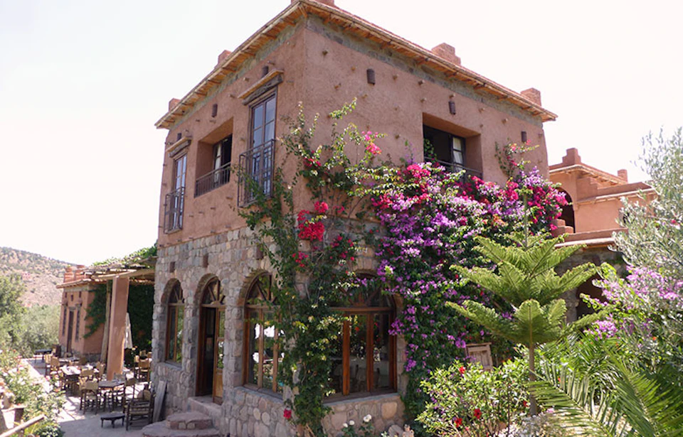 Two-story rustic stone and stucco house with flowering vines covering one side, arched windows, and a small balcony.