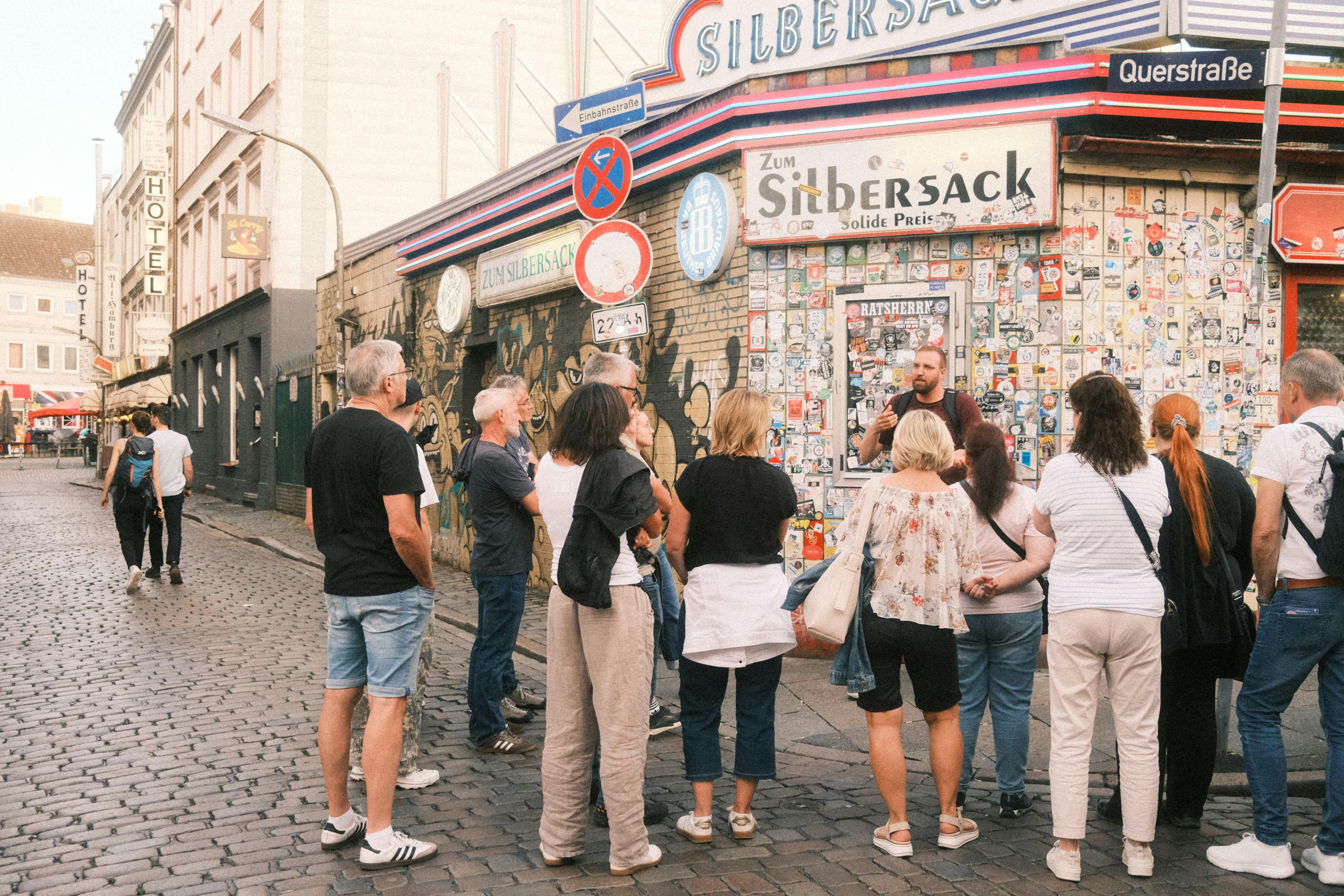 Μια ομάδα ανθρώπων συγκεντρώνονται μπροστά από ένα μπαρ καλυμμένο με αυτοκόλλητο με μια πινακίδα που γράφει "Zum Silbersack". Η σκηνή διαδραματίζεται σε έναν πλακόστρωτο δρόμο.