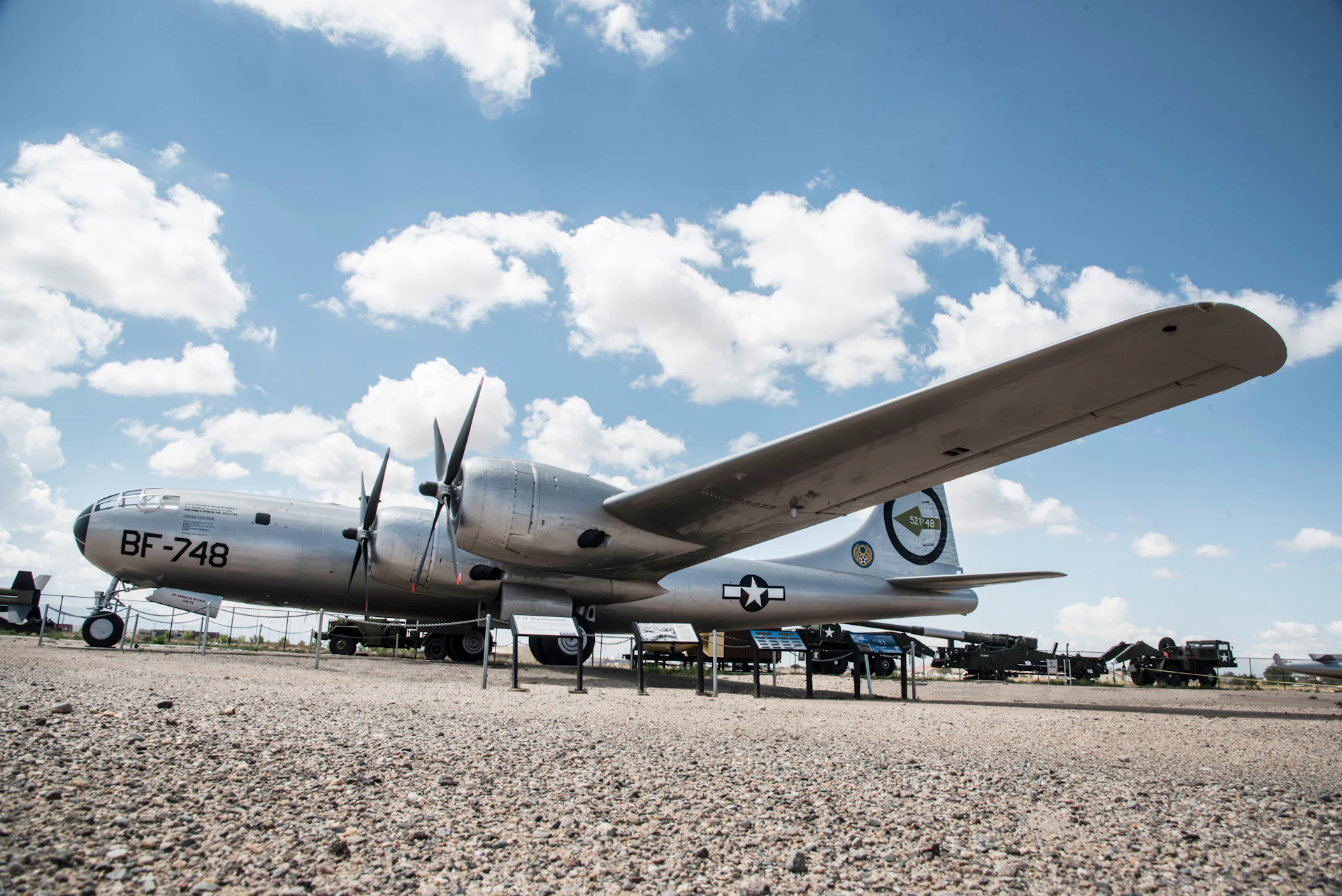 A silver World War II-era aircraft on display outdoors with propellers and military markings, under a partly cloudy sky.