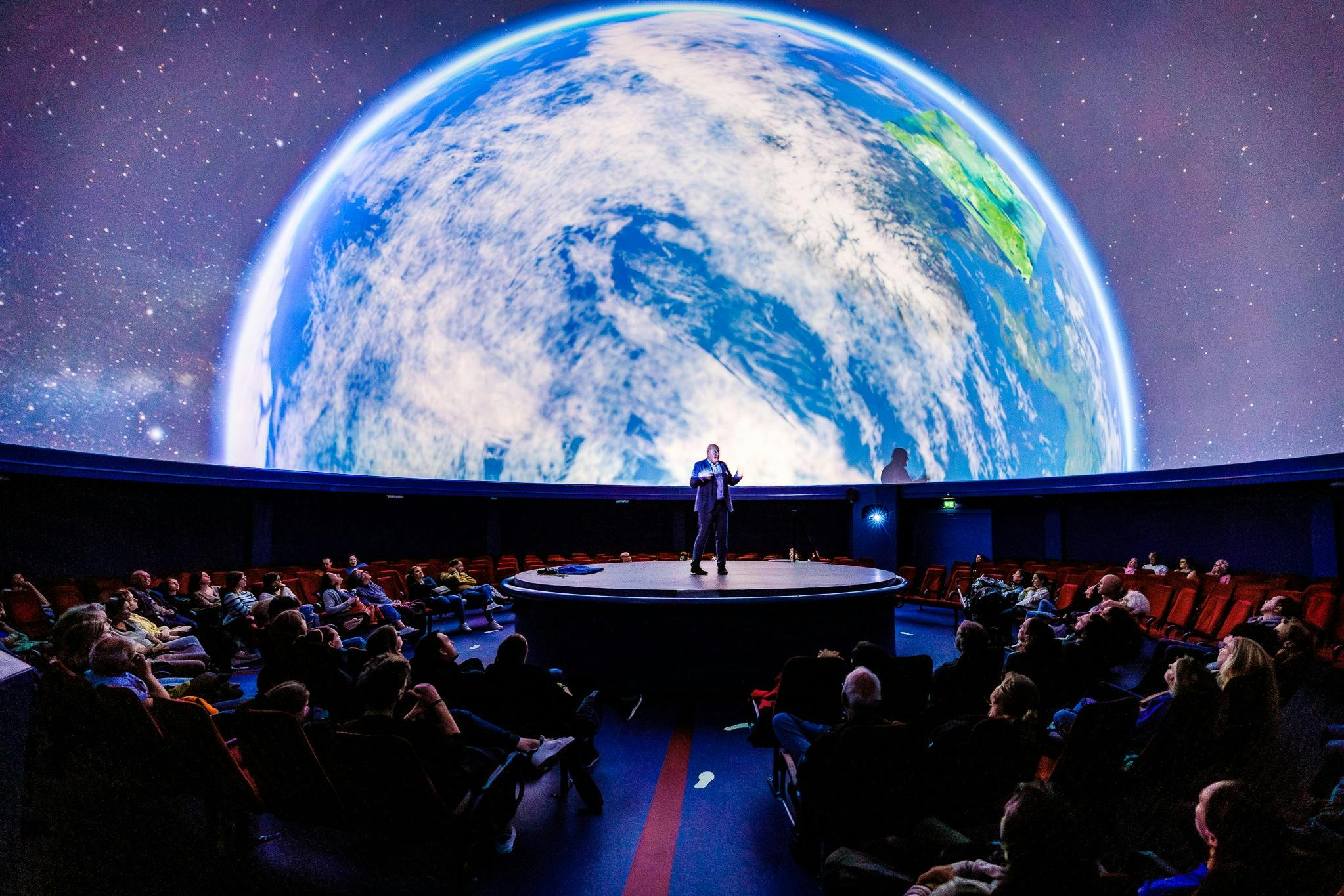 A presenter on a central stage in a theater with a large Earth projection on a dome screen above the audience.