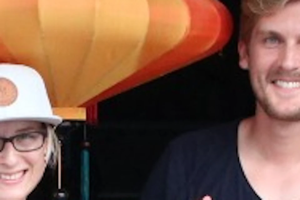 Two people smiling while standing in front of a lantern-making shop, holding colorful lanterns under a thatched roof.