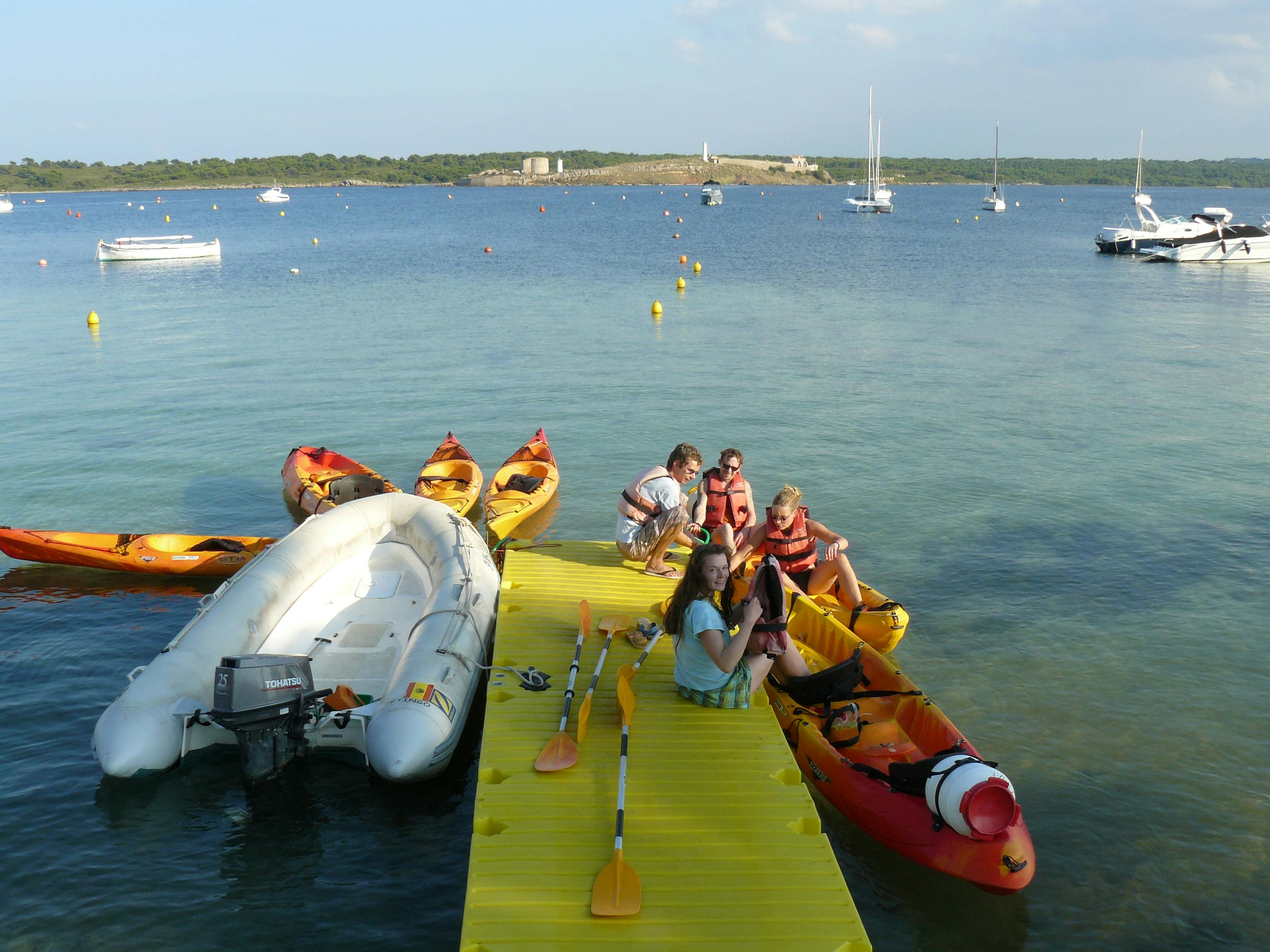 海のそばの黄色いドックに座っている人々は、遠くにブイとヨットがあり、カヤックと膨らませたボートに囲まれています。