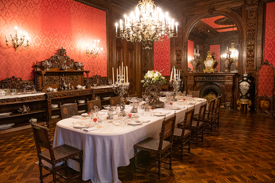 An ornate dining room with a long table set for a meal, surrounded by high-back chairs, chandeliers, and rich decorative elements.
