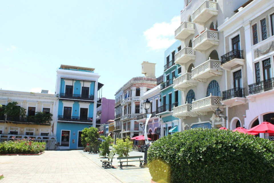 A sunny street lined with colorful colonial-style buildings and some trees, benches, and umbrellas in the foreground.