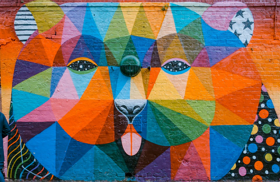 Colorful mural on a brick wall depicting a bear's face using geometric shapes and vibrant shades of blue, orange, green, and pink.