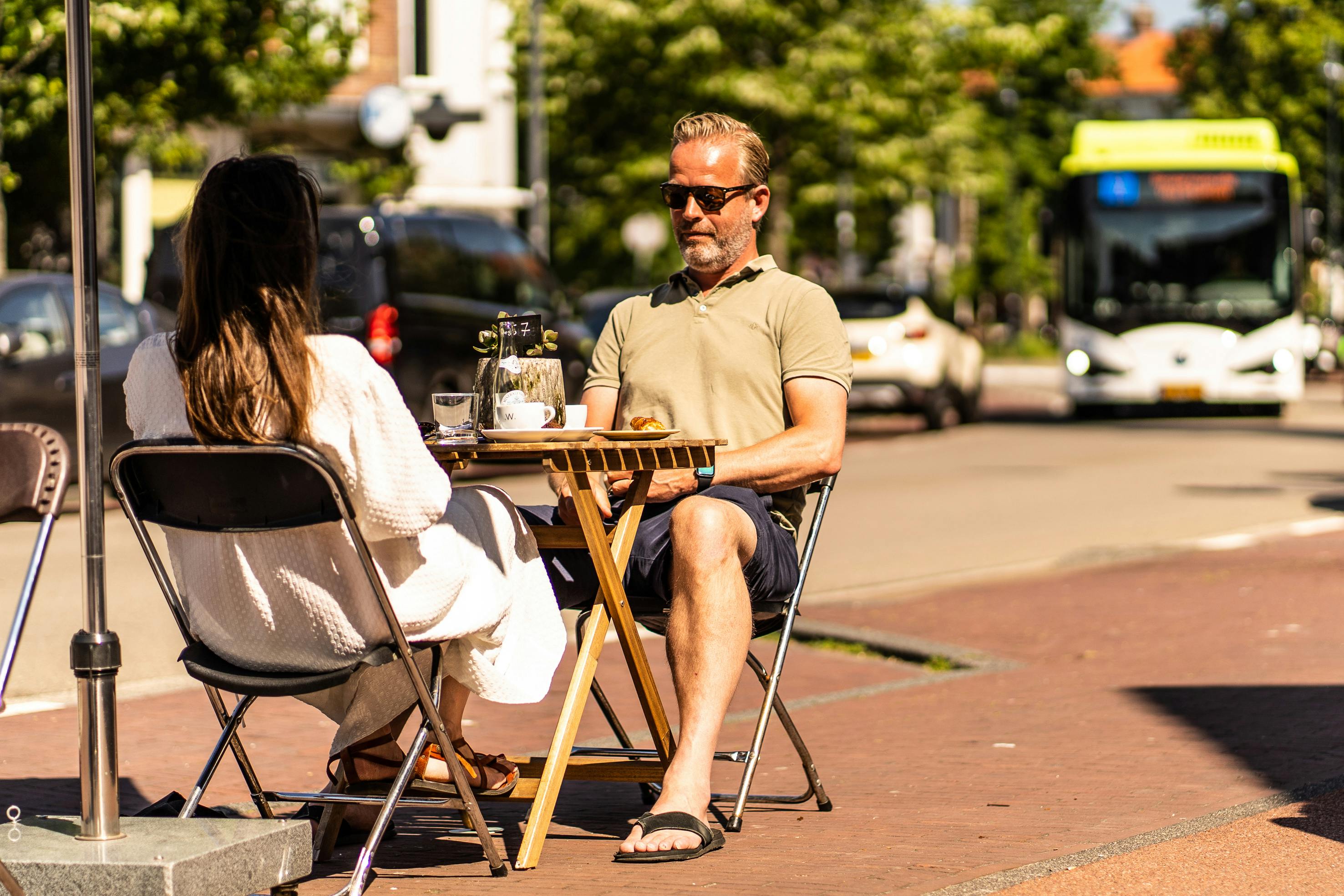 Δύο άνθρωποι κάθονται σε ένα υπαίθριο τραπέζι καφέ, ο ένας απέναντι από την κάμερα, με το φως του ήλιου και μια σκηνή του δρόμου στο παρασκήνιο.