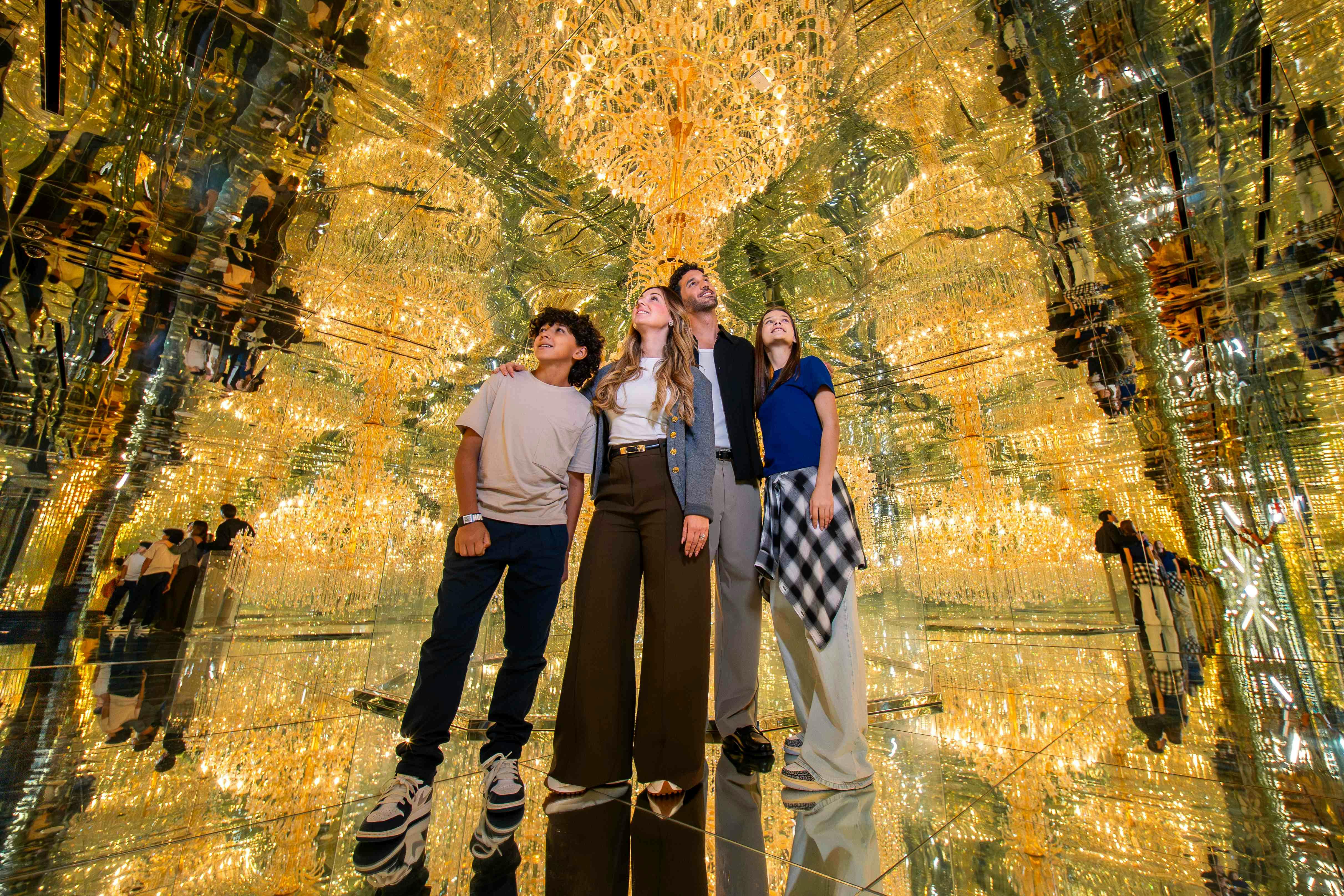 Four people standing together in a room with mirrored walls and ceilings, surrounded by glowing golden lights in a reflective setting.