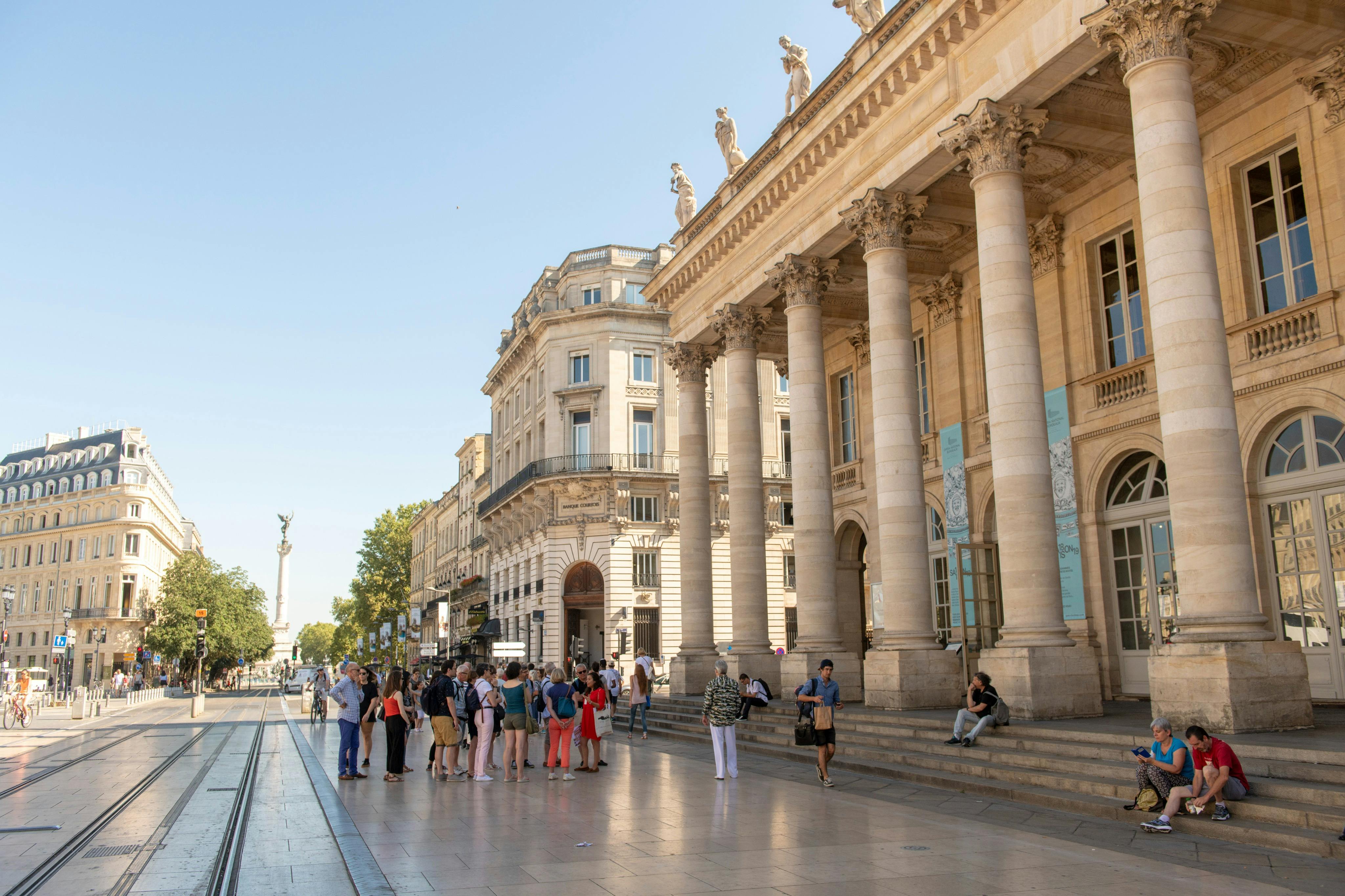 En grupp människor står utanför en storslagen byggnad med pelare och statyer på taket, med en klarblå himmel ovanför.