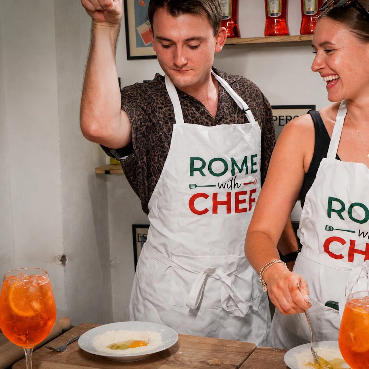 Two individuals in aprons, labeled "Rome with Chef," preparing dishes at a counter with drinks in the foreground.