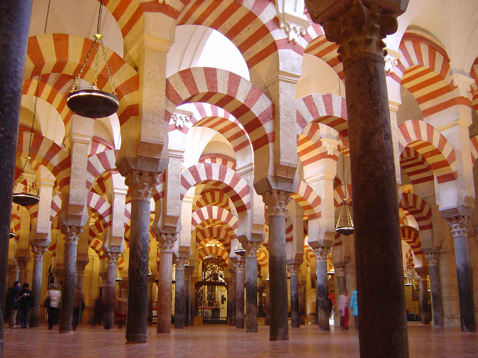 A vast hall with ornate red and white striped arches supported by stone pillars, featuring chandeliers and patterned tile floor.