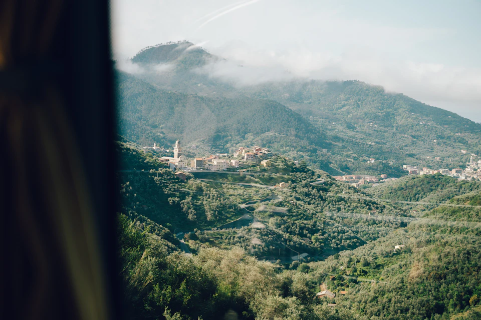 Aerial view of a small village with a prominent tower nestled in lush green hills and valleys under a partly cloudy sky.