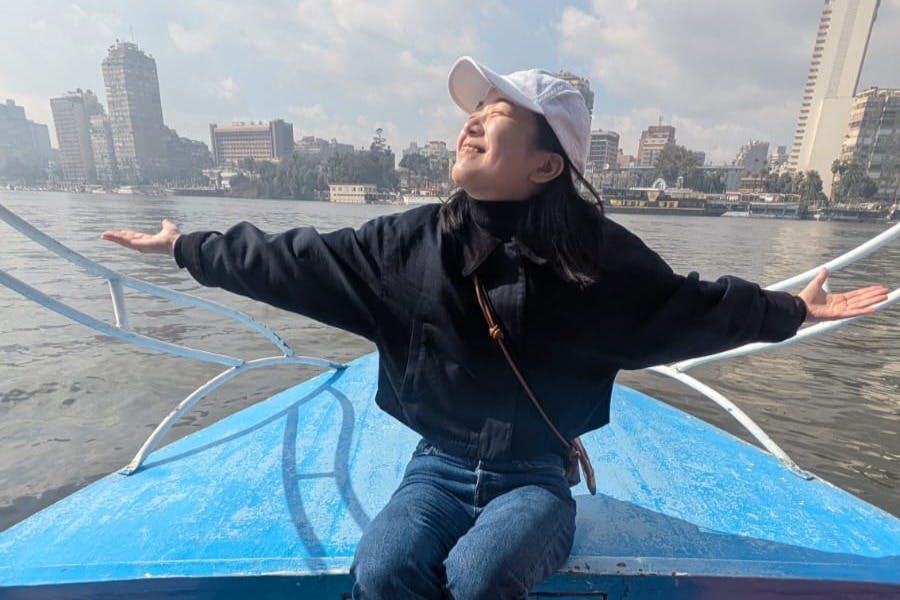 Ένα άτομο κάθεται σε μια βάρκα, χαμογελώντας με τα χέρια τεντωμένα, το αστικό τοπίο και το ποτάμι στο παρασκήνιο κάτω από έναν εν μέρει συννεφιασμένο ουρανό.